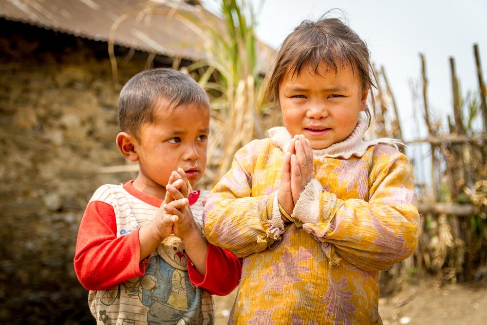 Children greeting with a traditional Namaste pose in a rural setting.