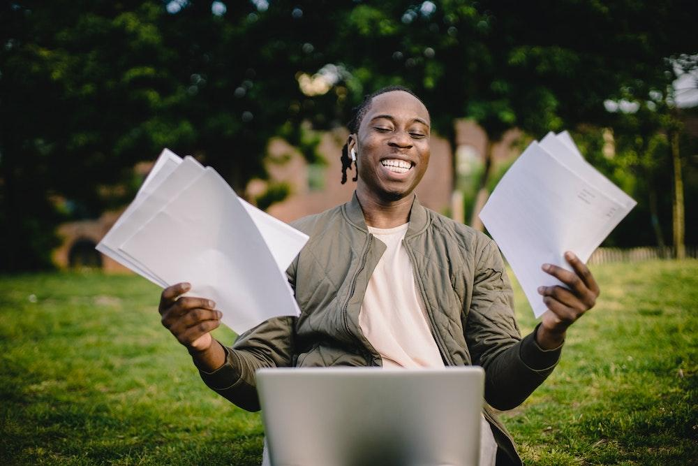 Happy student celebrating success with papers and a laptop outdoors.