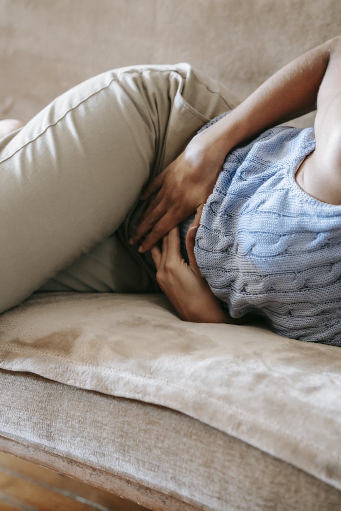 Woman in casual outfit resting on sofa, holding her stomach in discomfort.